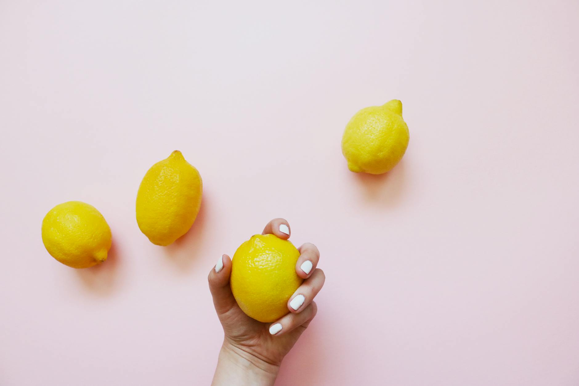 A hand holding a bright lemon on soft pink background with additional lemons arranged nearby