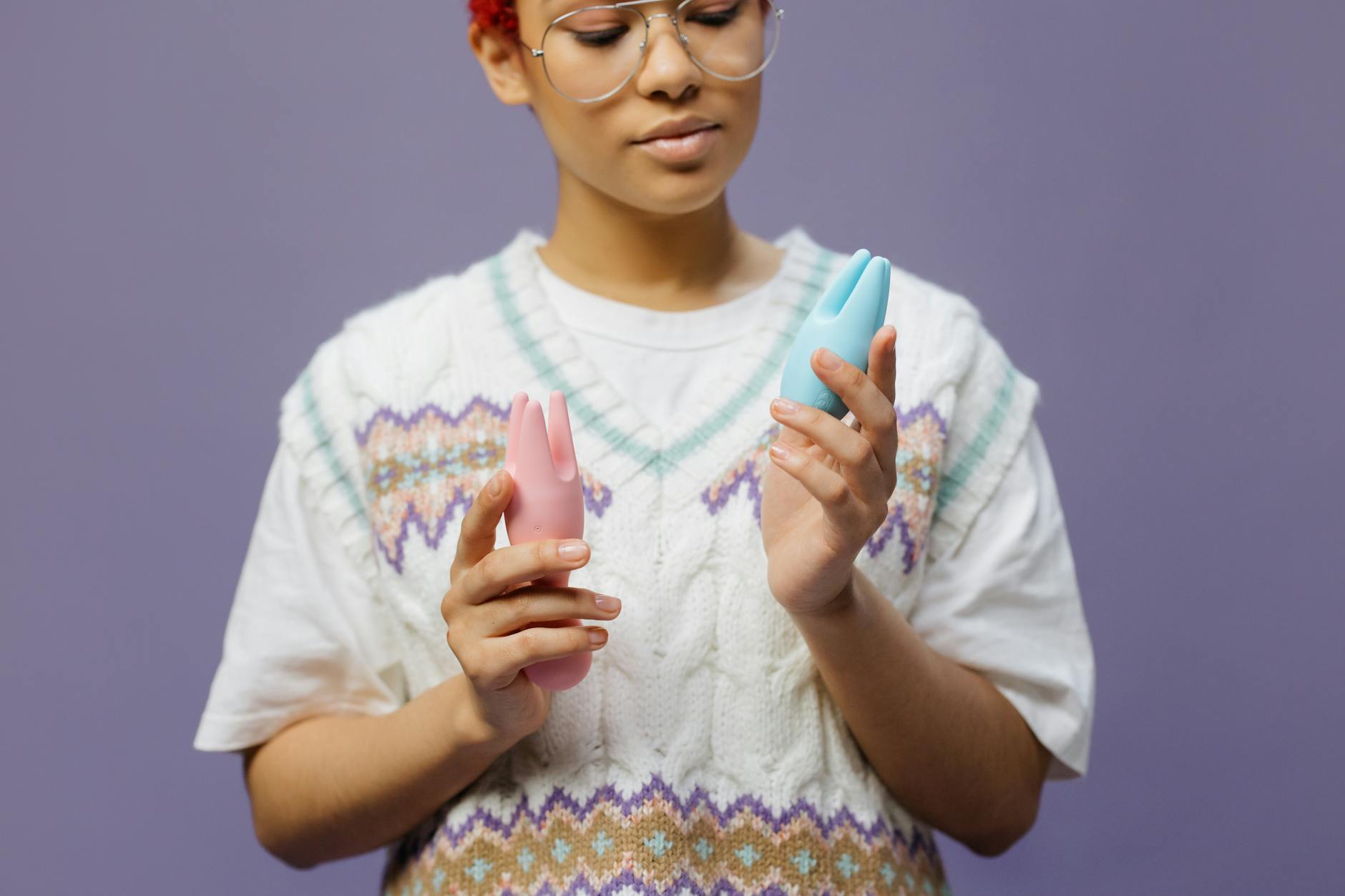 Woman holding blue and pink vibrators, contemplative expression against neutral background